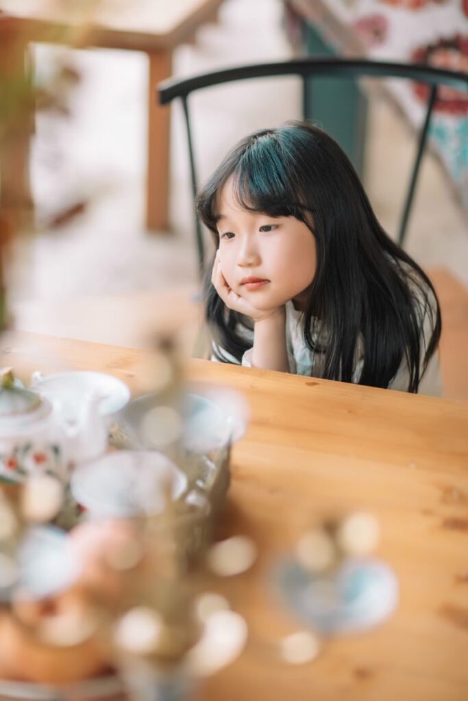 Young Asian girl with long black hair looking thoughtful while sitting at a wooden table indoors.