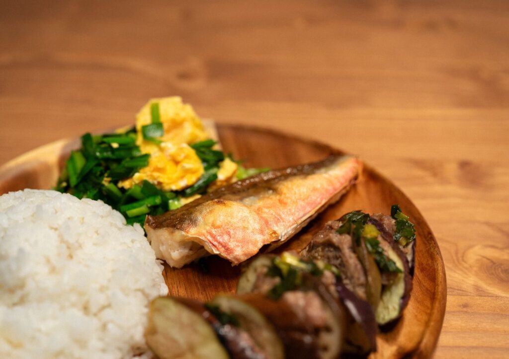 A wooden plate featuring white rice, grilled fish, sautéed vegetables, and scrambled eggs, showcasing Asian cuisine.