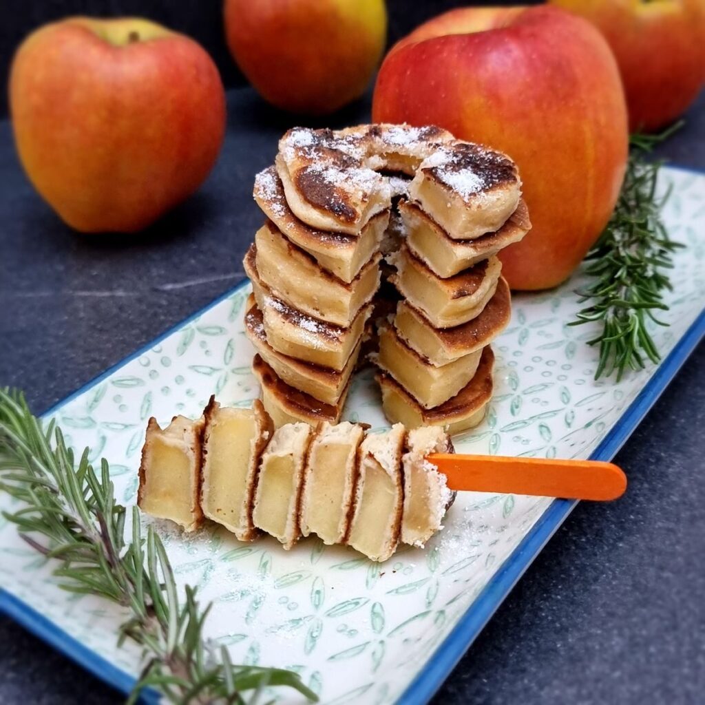 Stacked apple ring pancakes dusted with powdered sugar, garnished with fresh apples and rosemary sprigs.