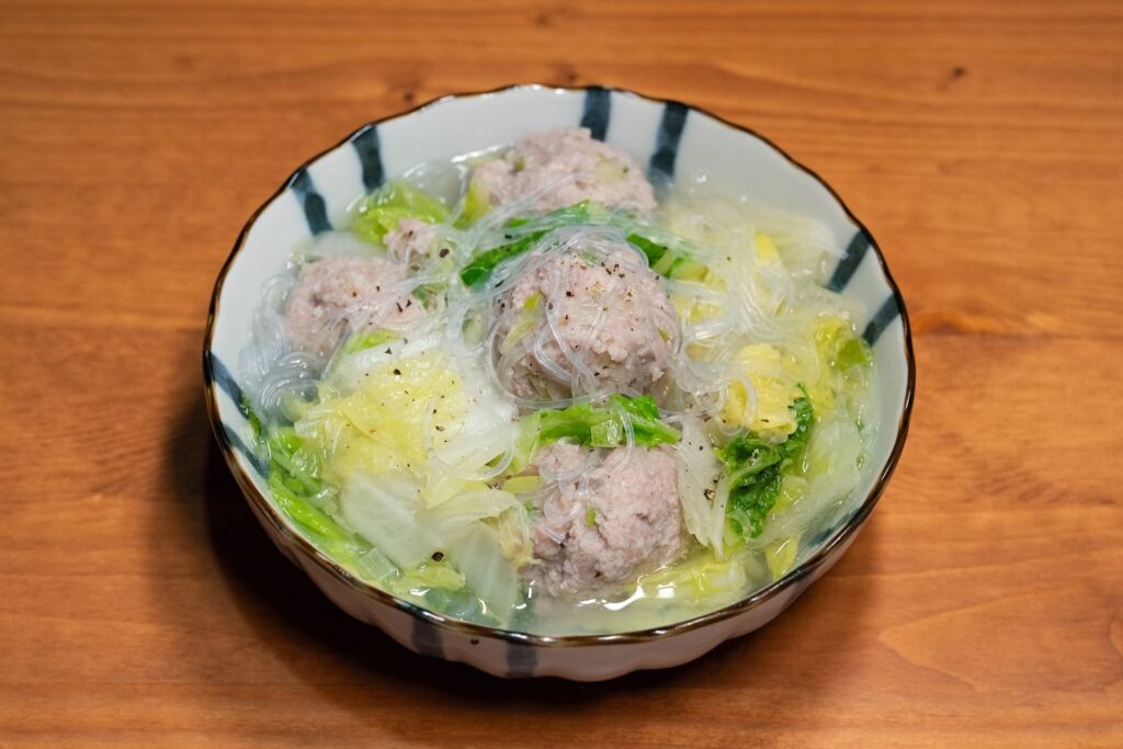Bowl of Asian meatball soup with glass noodles on a wooden table, showcasing vibrant vegetables.